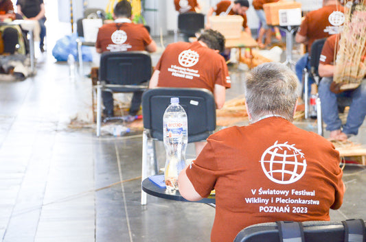 Alt（英語 / 日本語）  Basket makers seated and weaving during the live competition at the 5th World Wicker and Weaving Festival 2023 in Poznań, wearing brown event T-shirts / ポーランド・ポズナンで開催された第5回世界かご編み大会2023の実演会場で、茶色の大会Tシャツを着てかごを編む参加者たち