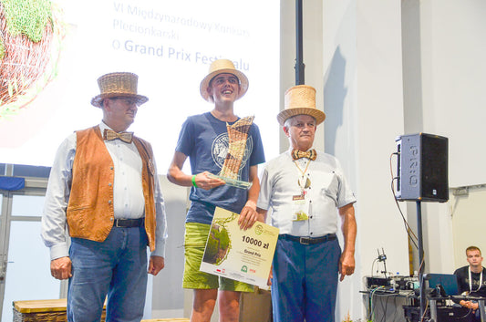 Grand Prix winner standing on stage with the woven basket trophy and certificate, flanked by two organizers at the World Wicker and Weaving Festival 2023 in Poland / 世界かご編み大会2023 in ポーランドにて、かごで編まれたトロフィーと賞状を手に、主催者に挟まれて立つ総合グランプリ受賞者