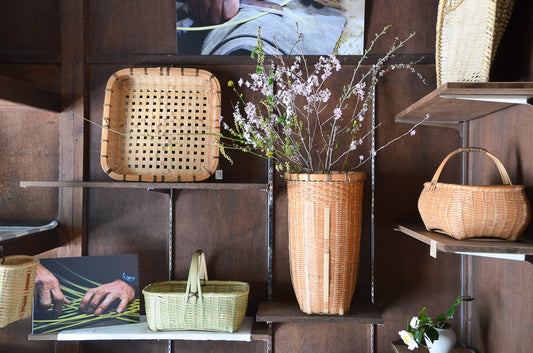 はい、承知しました。 **SP用 Alt（英語 / 日本語）**で出します。写真に忠実・簡潔です。  Alt： Exhibition display of bamboo baskets and trays by Hiromi Kiriyama, including a tall flower basket with branches / 企画展会場に並ぶ桐山浩実さん制作の竹籠と折敷、枝物を生けた背の高い花籠の展示