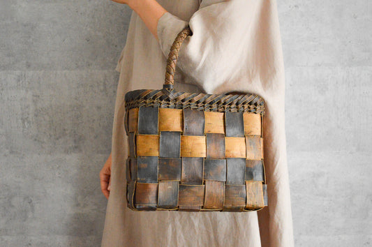 了解です。スラッシュ区切り・一文で出します。  Alt Handheld sawagurumi (walnut bark) basket bag with a checkered weave, carried by a person wearing a light linen dress / リネンの服を着た人物が、さわぐるみ（くるみ樹皮）で編まれた市松模様のかごバッグを手に持っている様子
