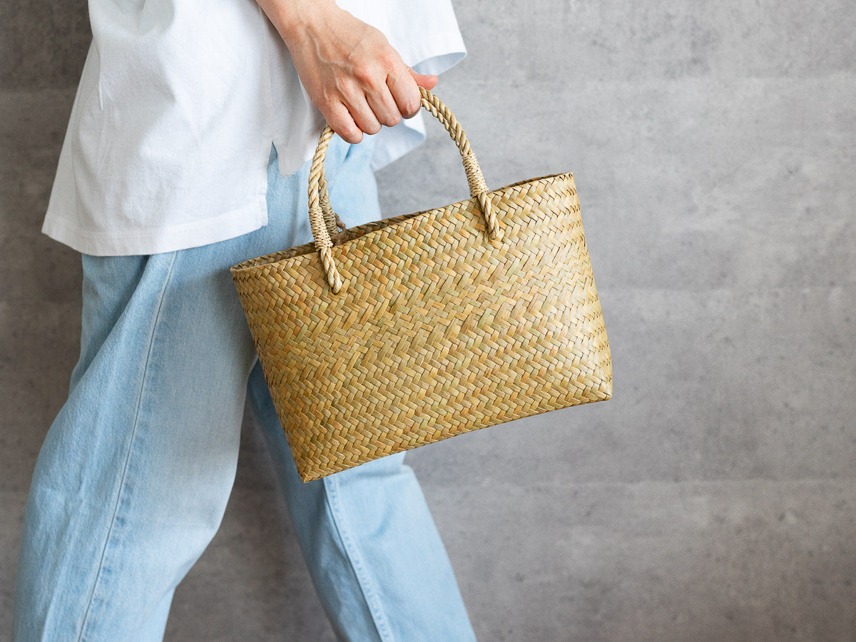 Hand holding a slim oval Krajood grass basket bag / カチューのスリムオーバルかごバッグを手に持った様子