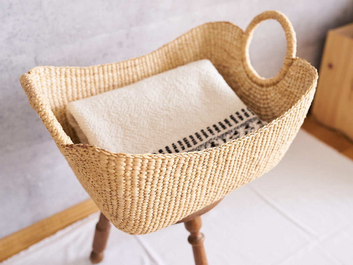 A water hyacinth organizing basket with a ring handle holding folded fabric on a wooden stool / リング状の持ち手が付いたウォーターヒヤシンスの整理かごに、折りたたんだ布を入れて木製のスツールに置いた様子